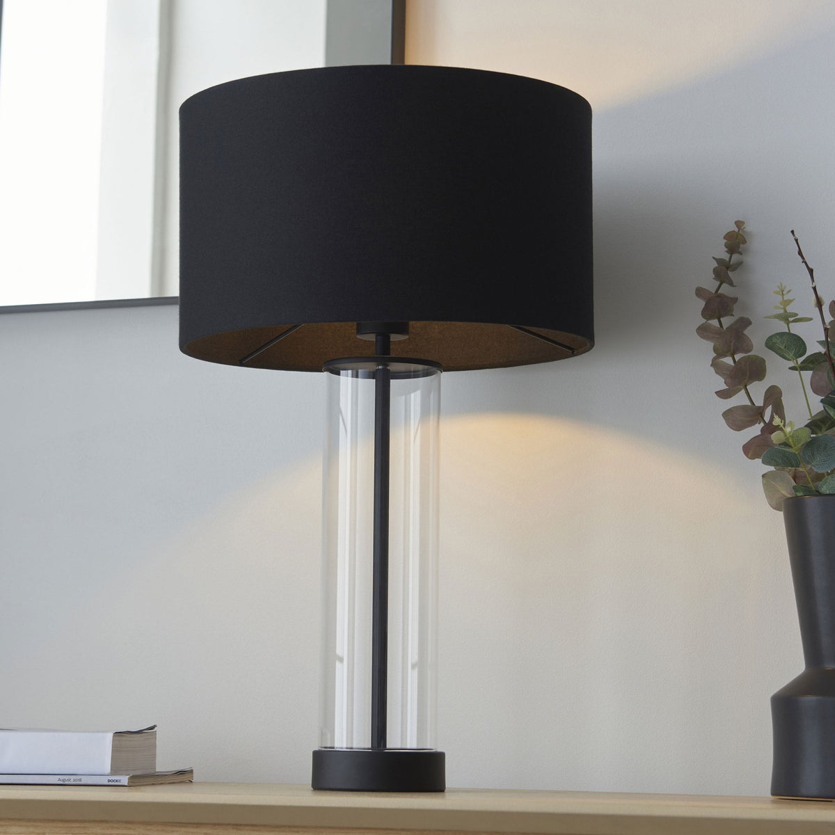 A black table lamp with a clear glass base stands on a light wooden surface in a contemporary room.