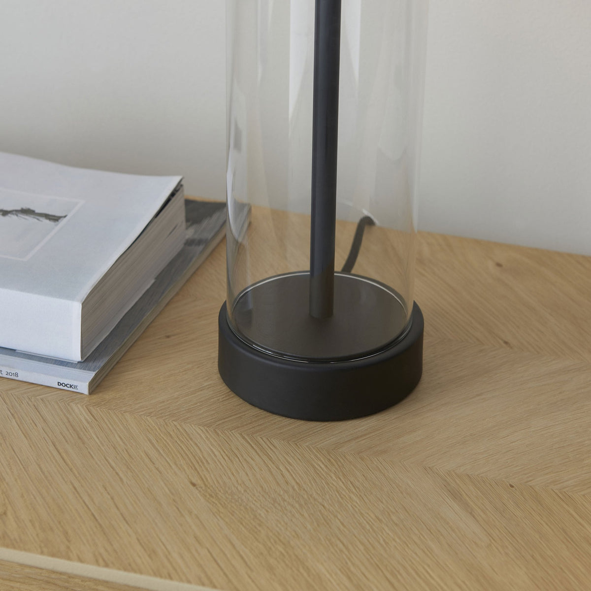 Close-up of the lamp's base, showing the clear glass cylinder meeting the black metal bottom on a wooden surface.