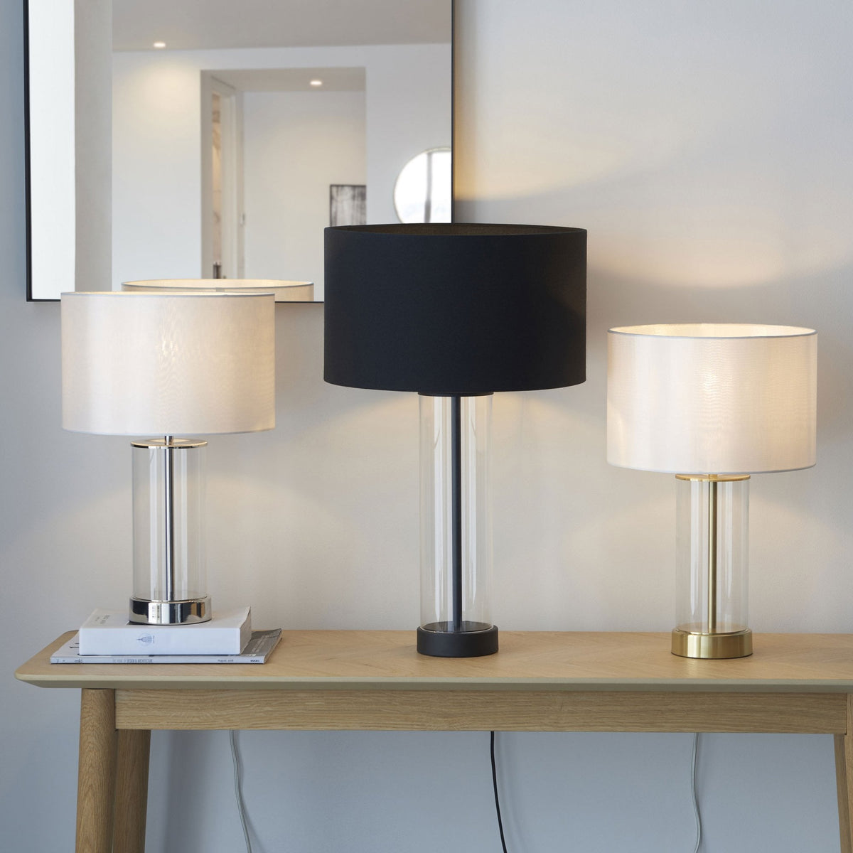 Three stylish table lamps with clear glass bases and different colored shades on a wooden console table.