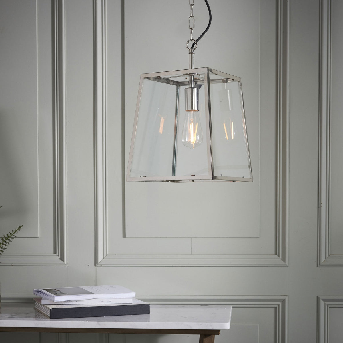 A glass and metal pendant light hangs above a table with books, against a light grey paneled wall.