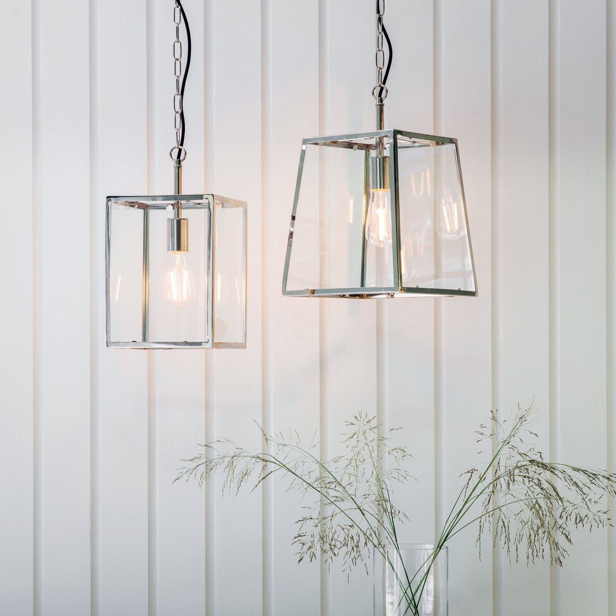 Two glass and metal pendant lights, one square and one tapered, hanging in a room with white paneling.