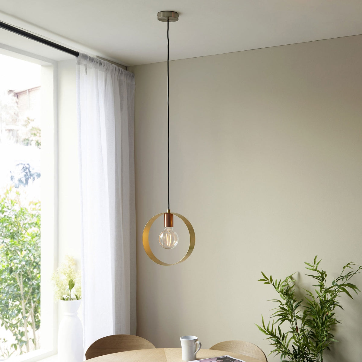 A pendant light with a brass ring and copper accent hangs above a light wood table in a dining room.