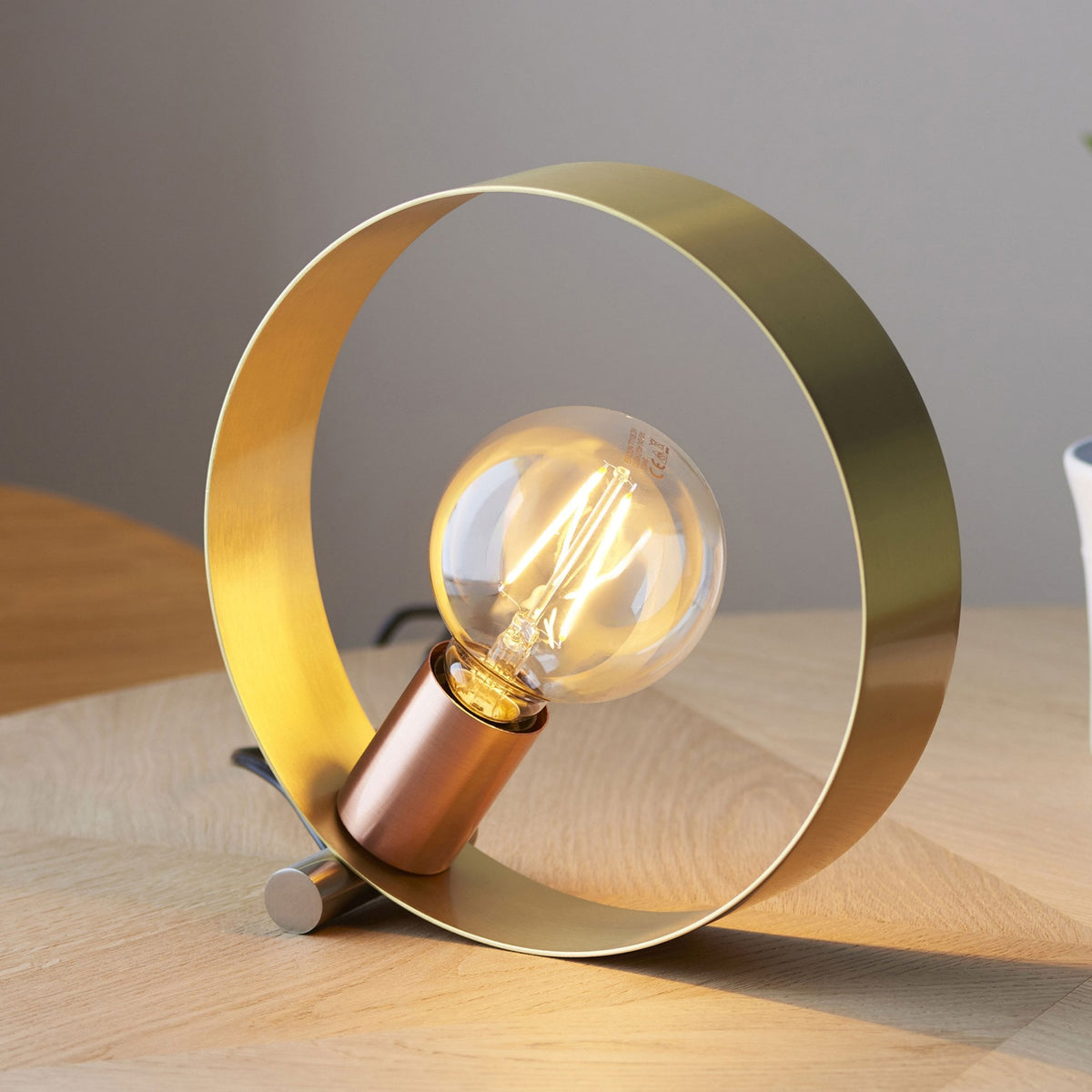 A close-up shot of a brass ring-shaped table lamp with a copper bulb holder and an exposed filament bulb.