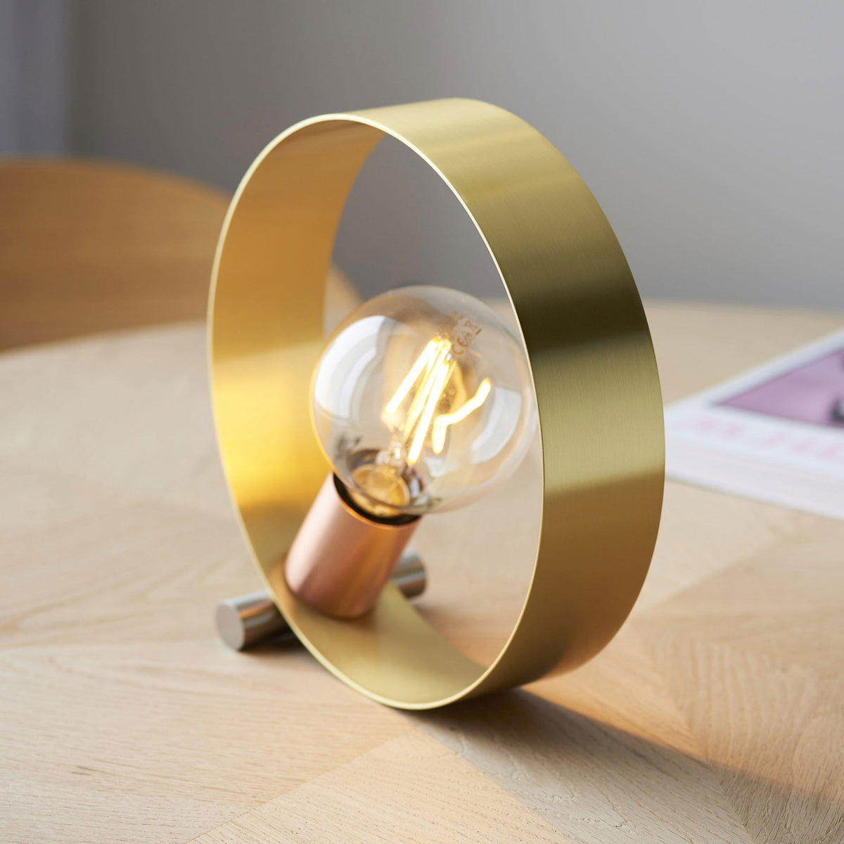 A brass ring-shaped table lamp with a copper bulb holder and an exposed filament bulb on a wooden table.