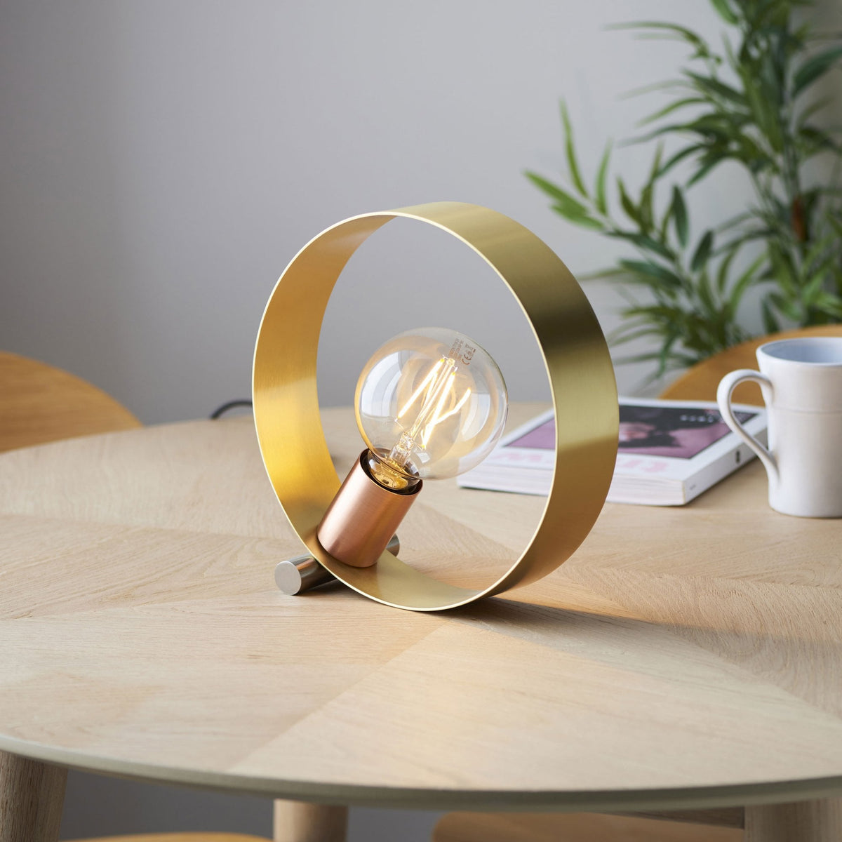 A brass ring table lamp with a glowing filament bulb sitting on a light wooden table, next to a white mug.