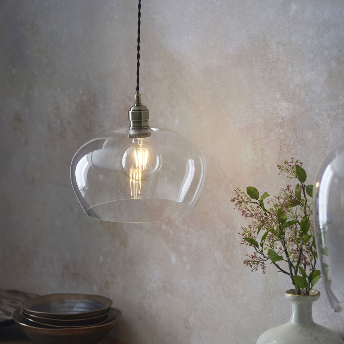 A clear glass pendant light with a visible filament bulb, hanging above a rustic wooden table with stacked plates.