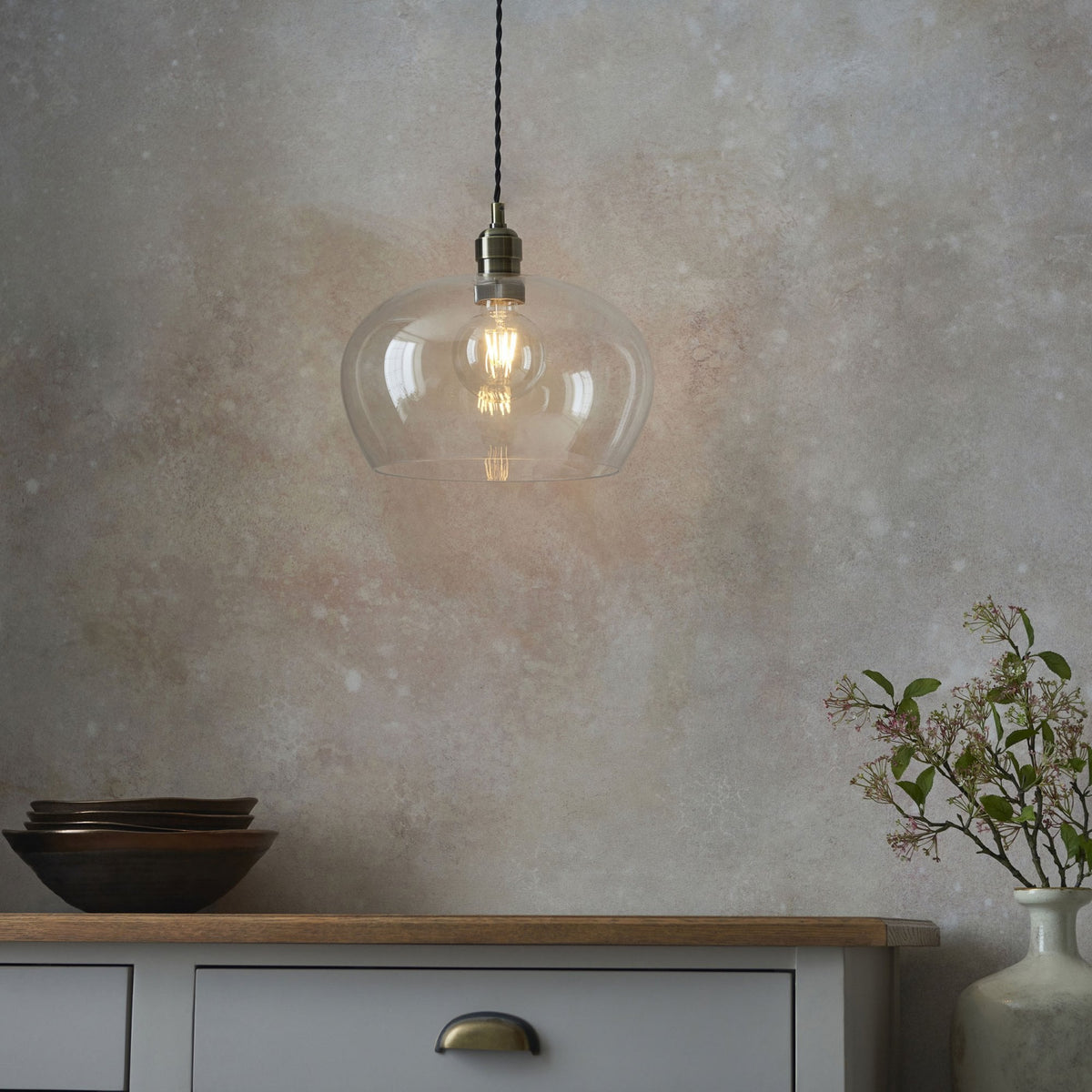 A clear glass pendant light hangs above a grey cabinet, illuminating the textured wall and a vase of flowers.