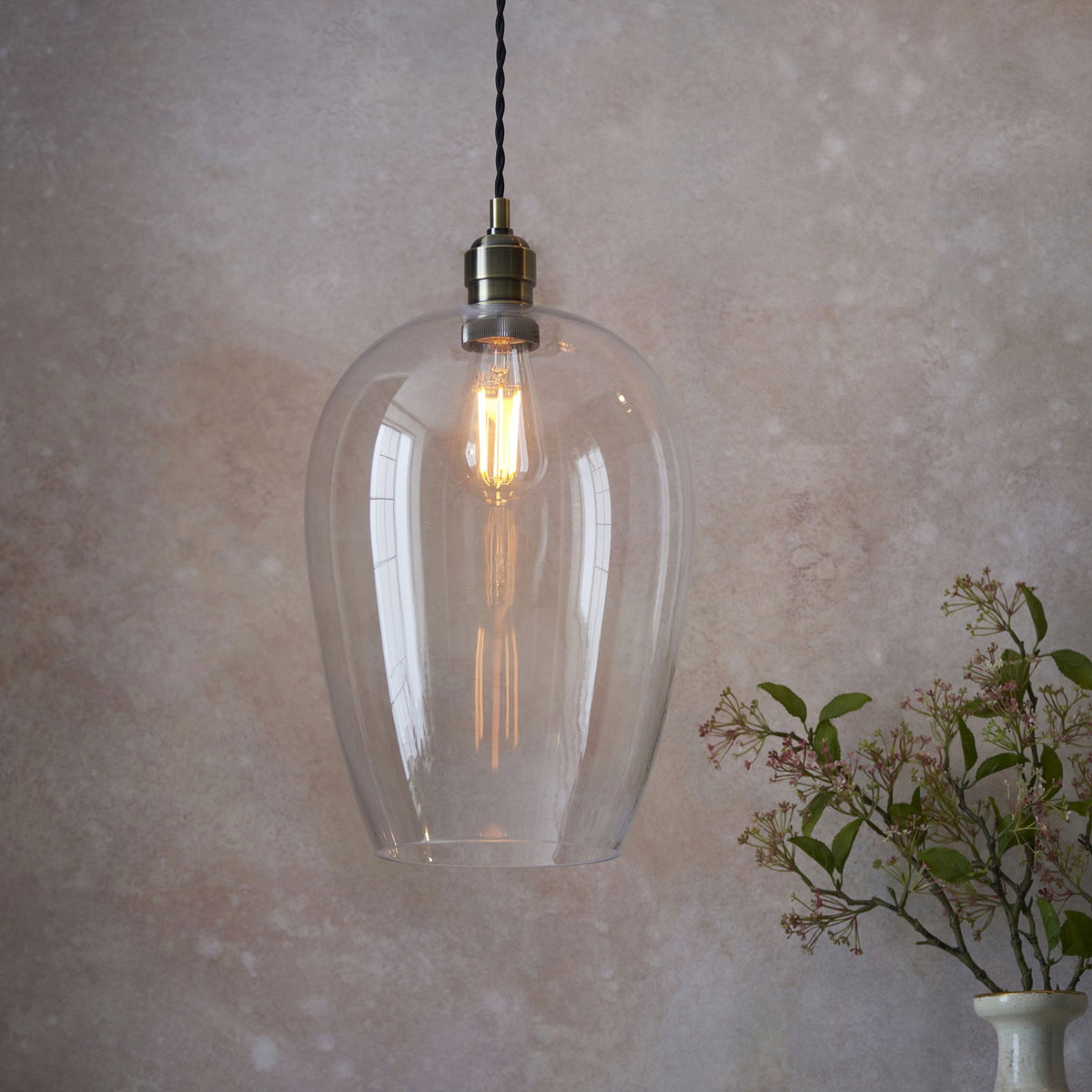A clear glass pendant light with an Edison bulb and brass fitting, hanging against a textured wall.