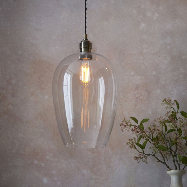 A clear glass pendant light with an Edison bulb and brass fitting, hanging against a textured wall.