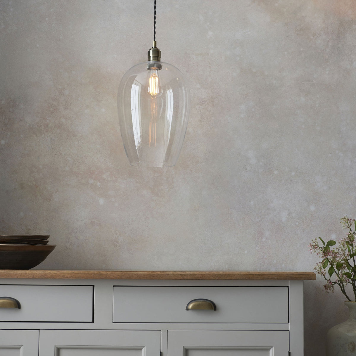 A clear glass pendant light illuminating a kitchen dresser with decorative bowls and flowers.