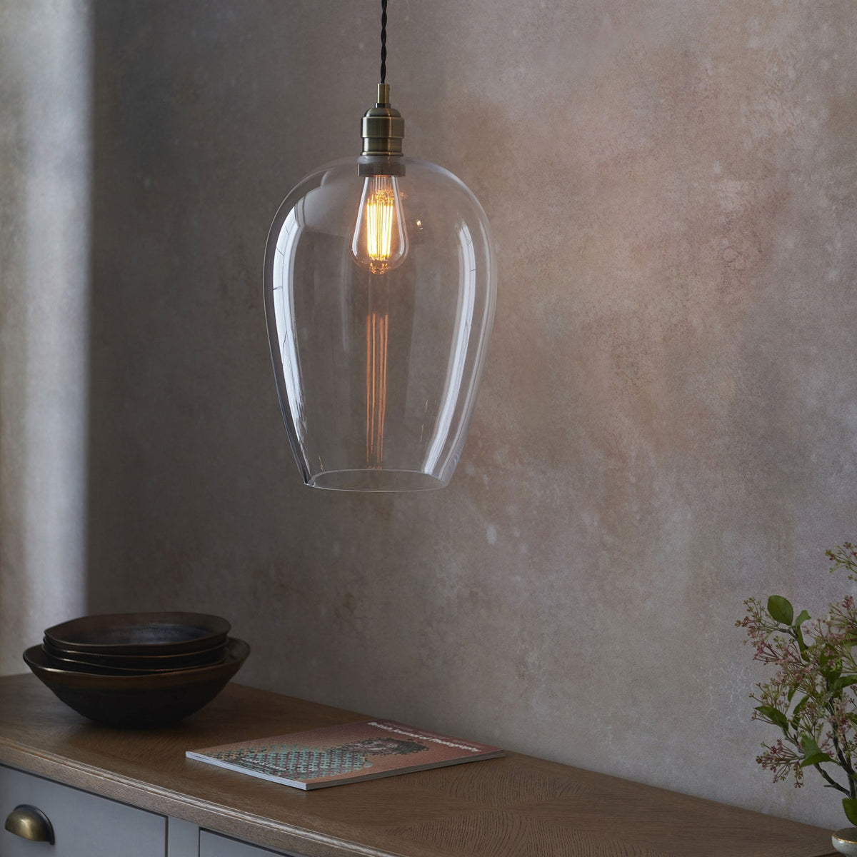 A clear glass pendant light casting a warm glow over a kitchen counter with bowls and a magazine.