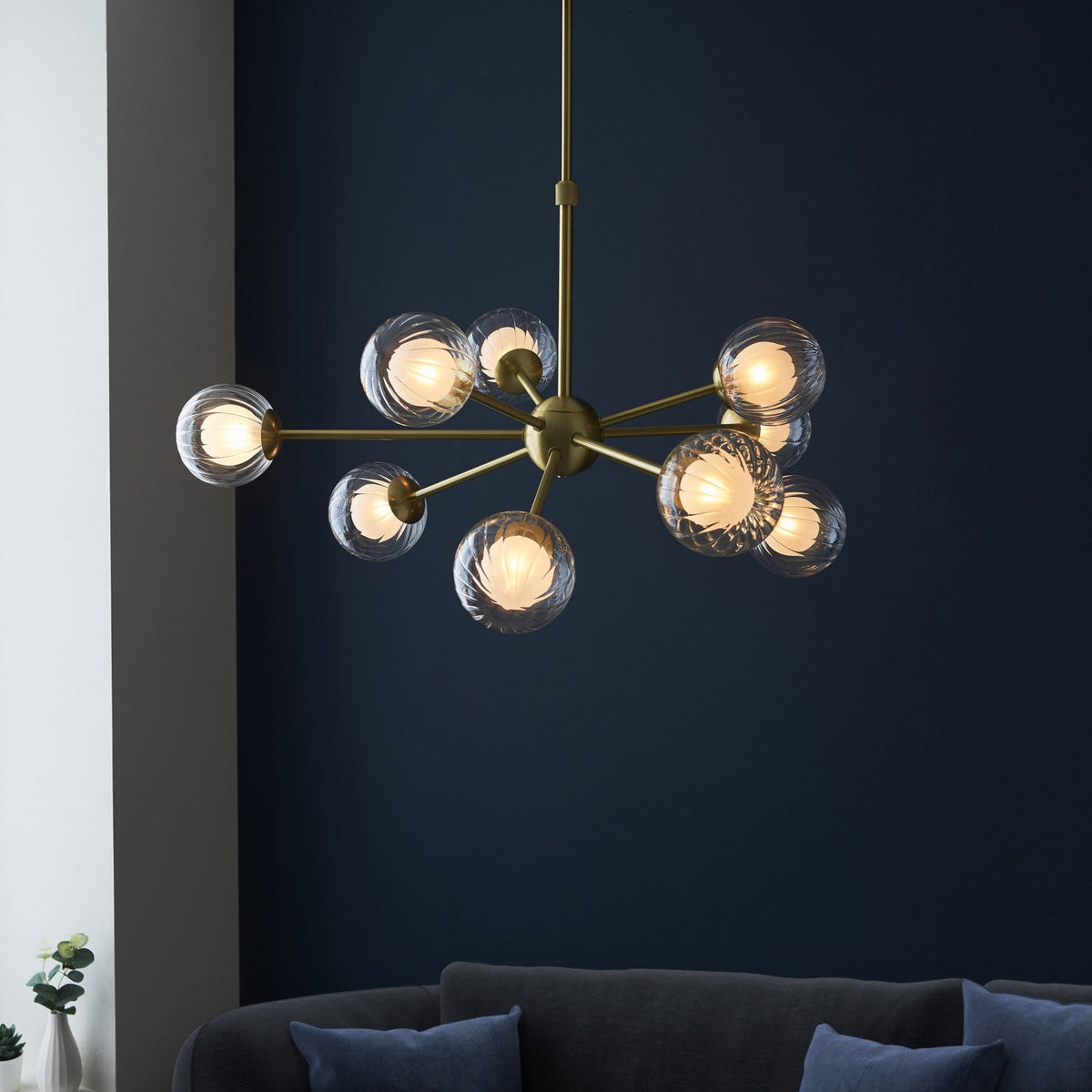 A modern brass chandelier with ten clear, textured glass globe lights hangs above a gray sofa.