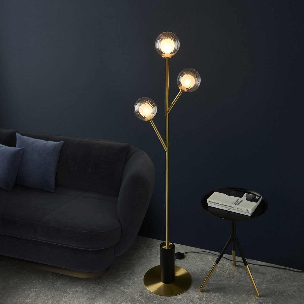 Lampadaire moderne en laiton et noir avec trois globes en verre texturé, éclairant un salon aux murs sombres.