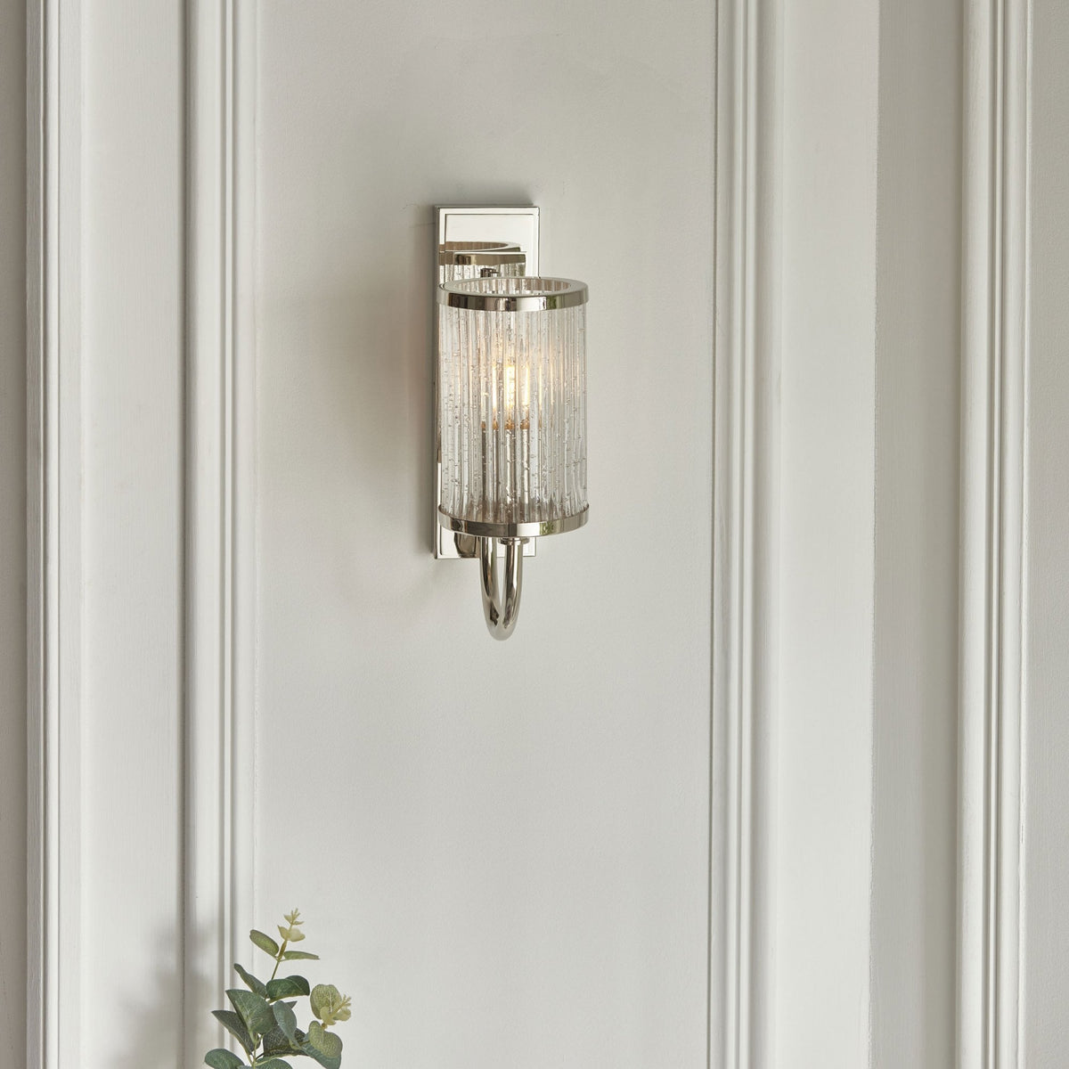 Un aplique de pared de cromo pulido con una pantalla de cristal acanalado burbujeante, iluminado, en un fondo de panel blanco.