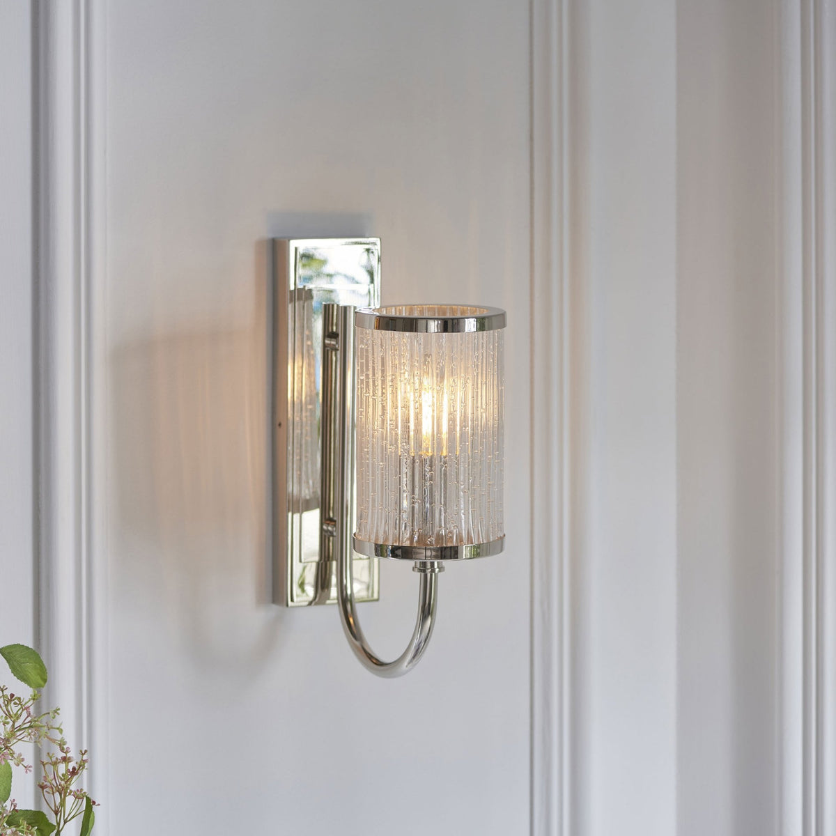 Un aplique de pared de cromo pulido con una pantalla de cristal acanalado burbujeante, iluminado, en un fondo de panel blanco.