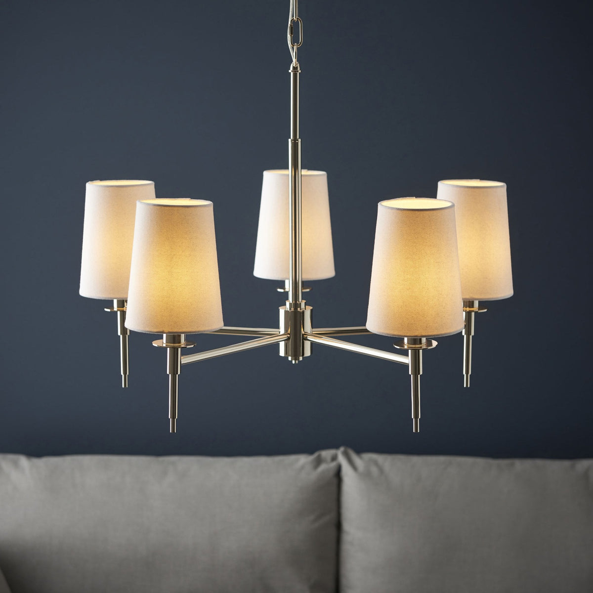 A modern 5-arm chandelier with cream fabric shades, glowing warmly against a dark blue wall.