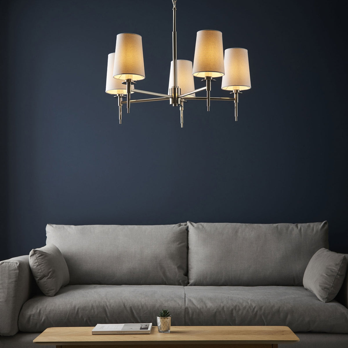 A modern 5-arm chandelier with cream shades illuminating a stylish living room with a grey sofa.