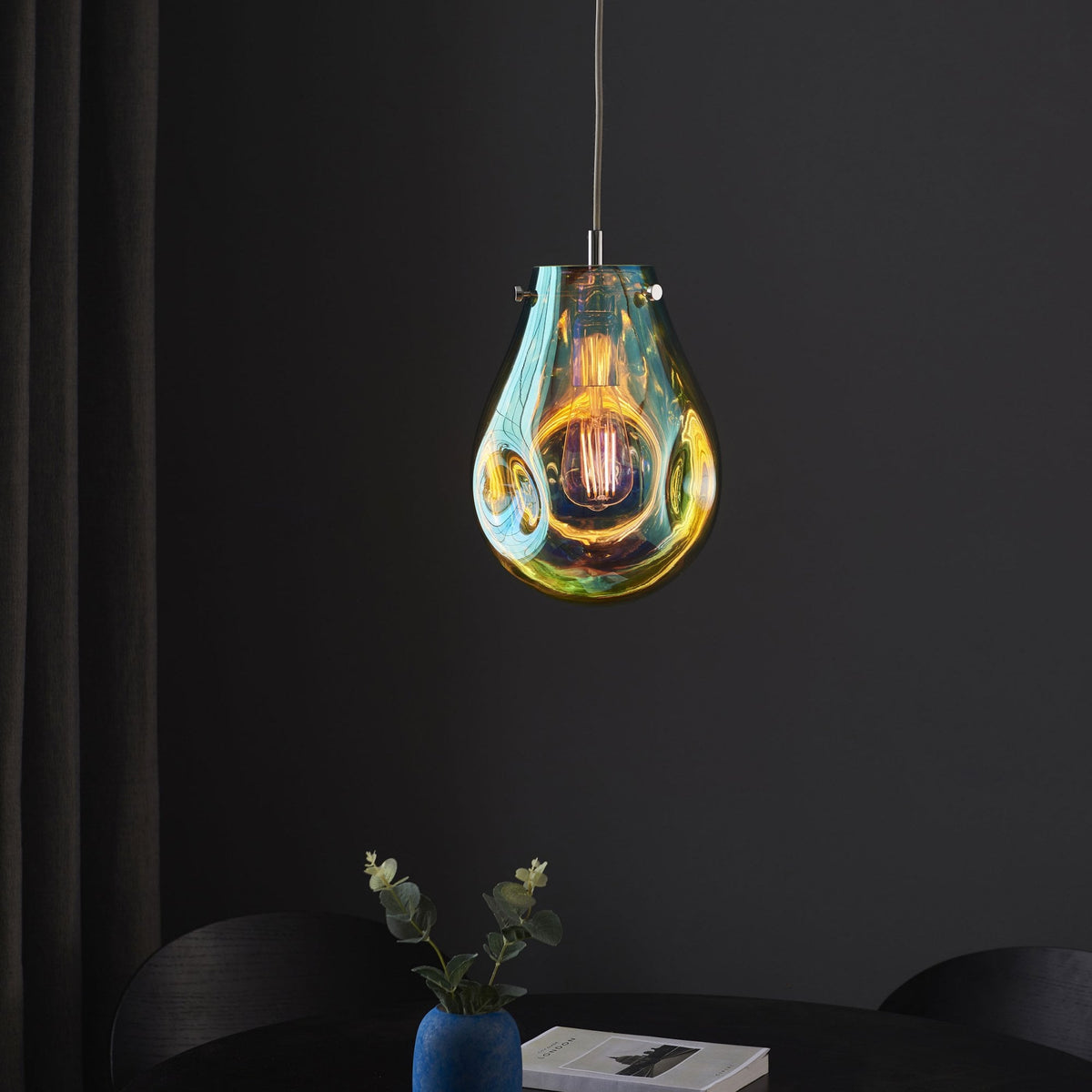 Teardrop pendant light in iridescent blue and amber with a lit bulb over a table.