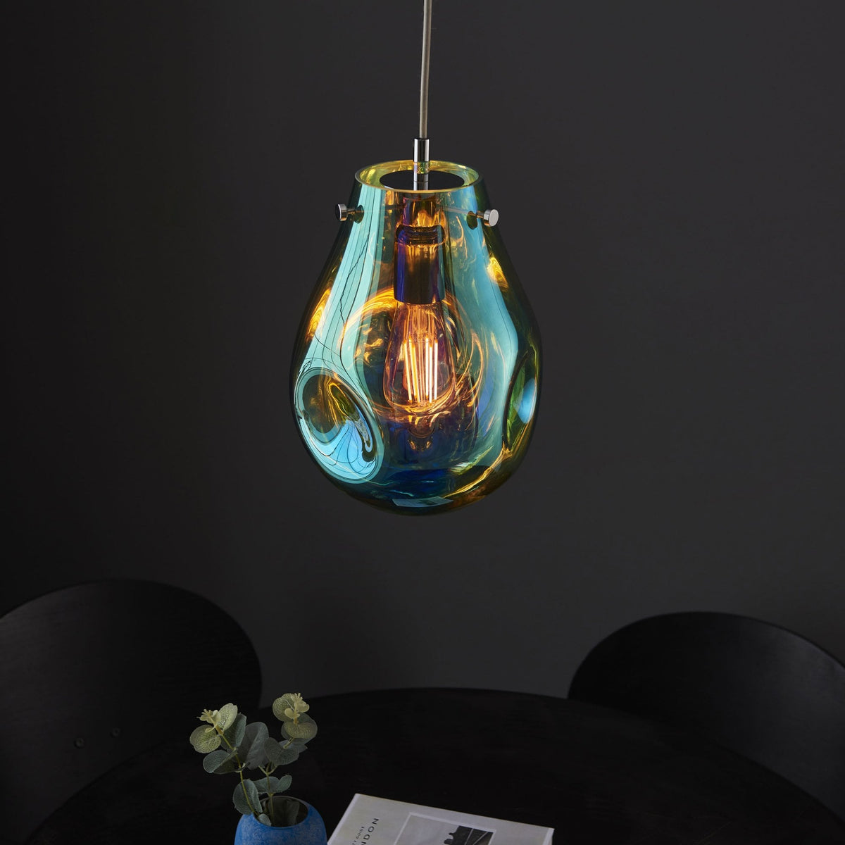 Teardrop pendant light in iridescent blue and amber with a lit bulb over a dark table.
