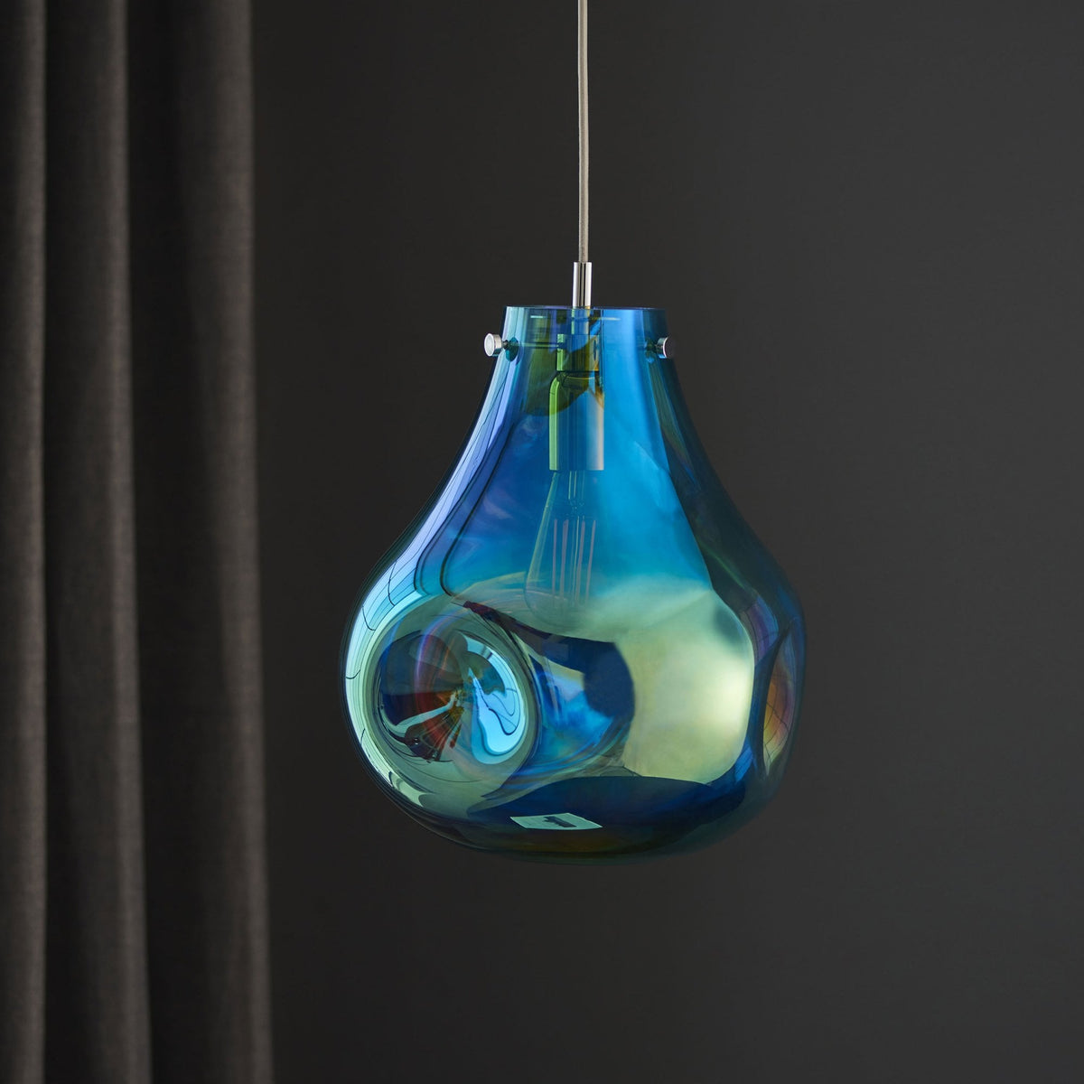 Iridescent glass pendant light, teardrop shape, with an unlit bulb, against a dark curtain.