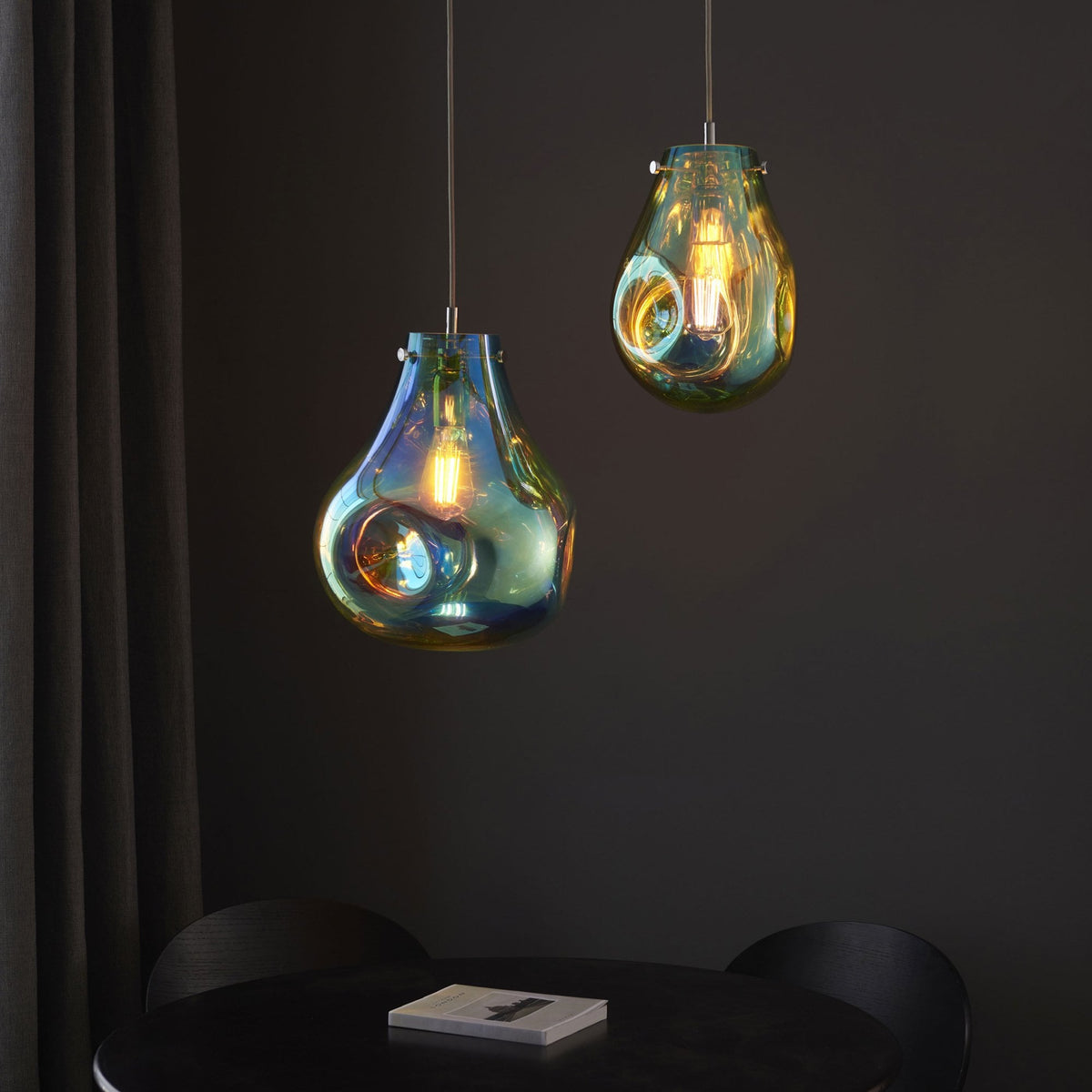 Two iridescent glass pendant lights hang over a dark dining table with a book.