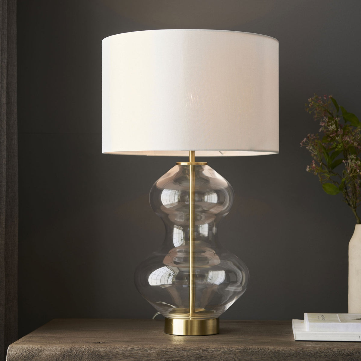A glass table lamp with a brass base and a white shade on a wooden table with a dark background.