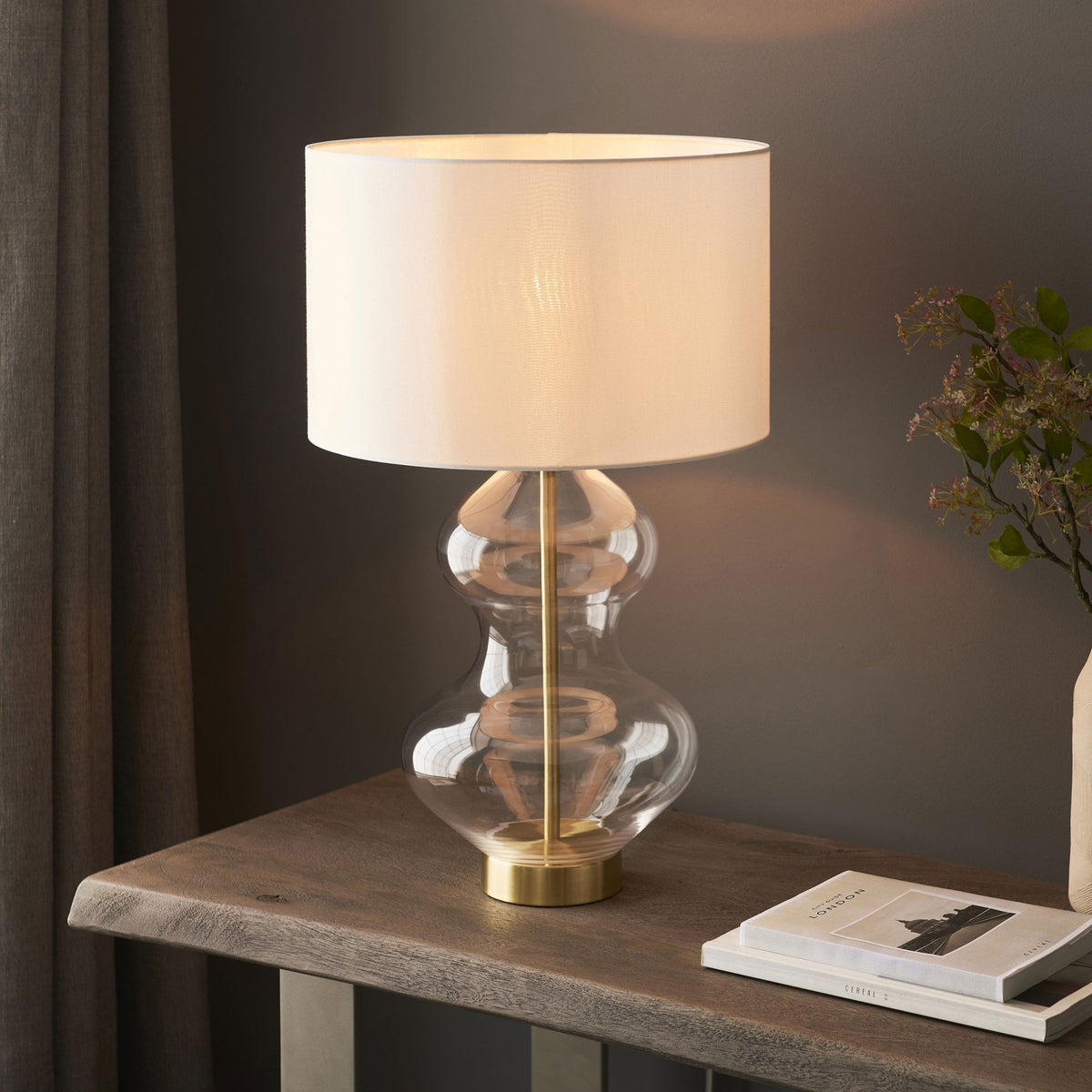 A glass table lamp with a brass base and a white shade, illuminated, on a wooden table.