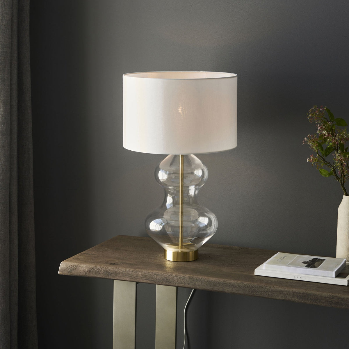 A glass table lamp with a brass base and a white shade, unlit, on a wooden console table.