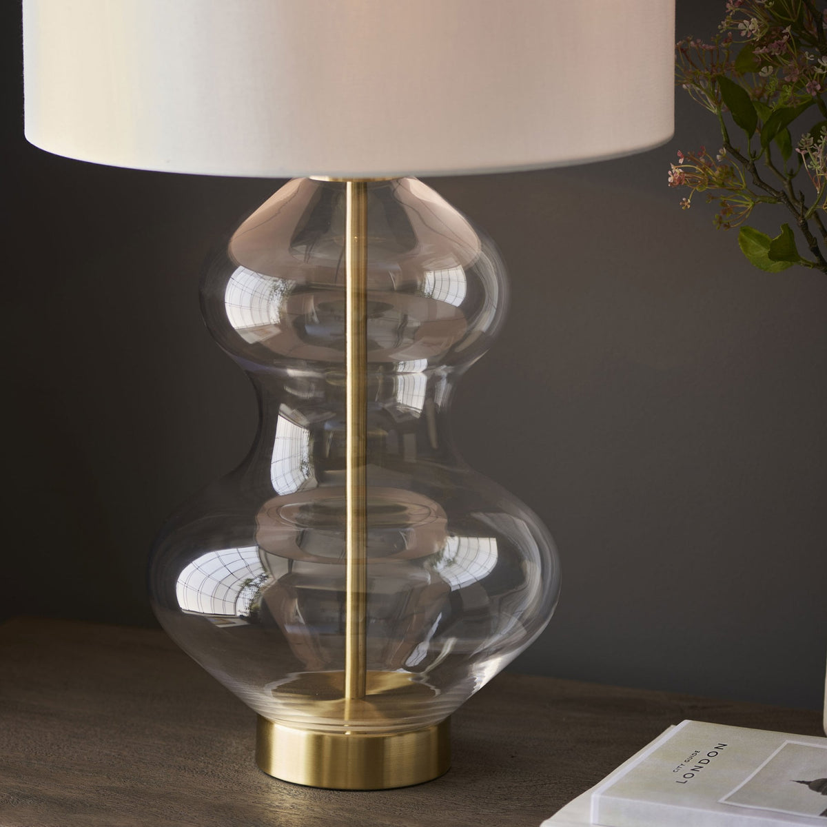 A close-up of a glass table lamp with a brass base and a white shade on a wooden table.