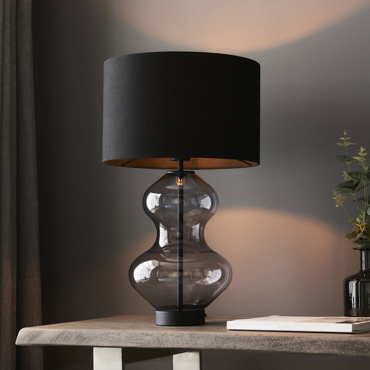 A modern table lamp with a dark gray hourglass-shaped glass base and a black cylindrical lampshade.
