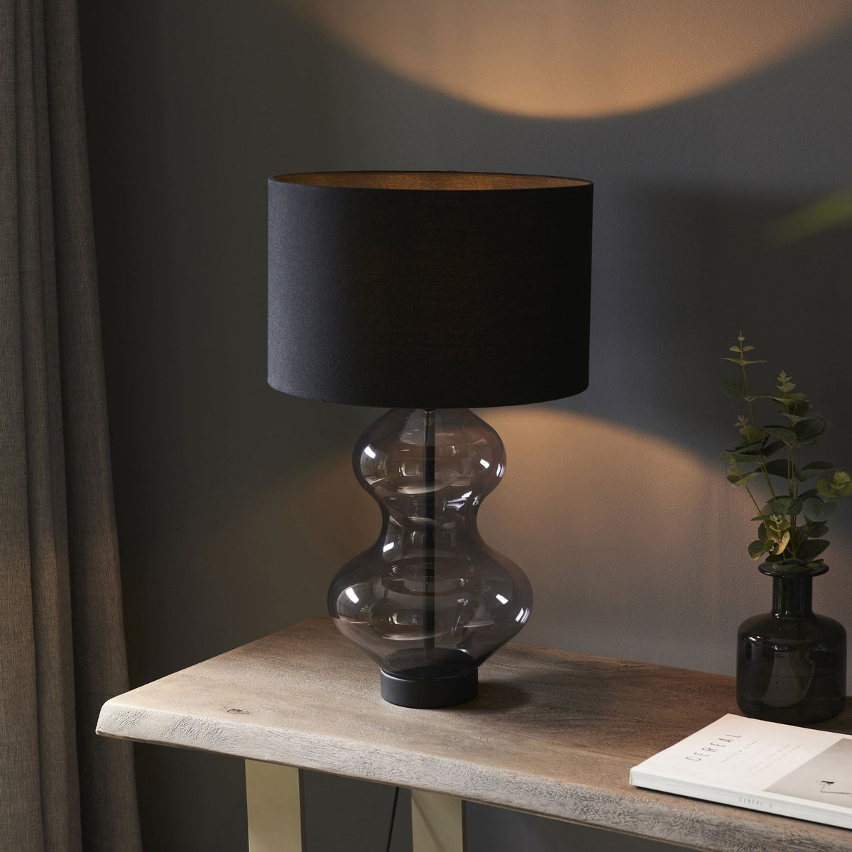 A modern table lamp with a dark gray hourglass-shaped glass base and a black cylindrical lampshade emitting warm light.