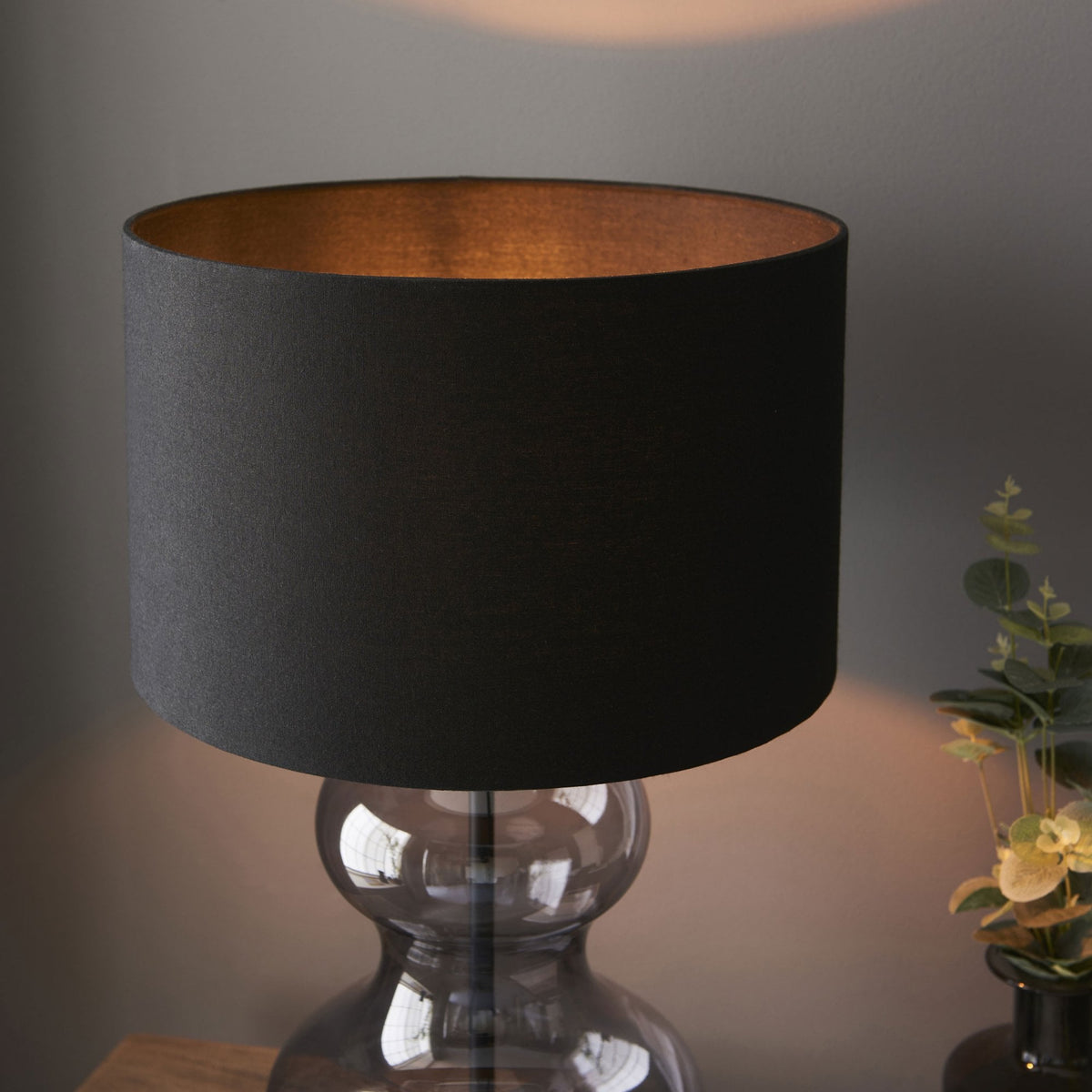 A close-up of a modern table lamp with a dark gray hourglass-shaped glass base and a black cylindrical lampshade.