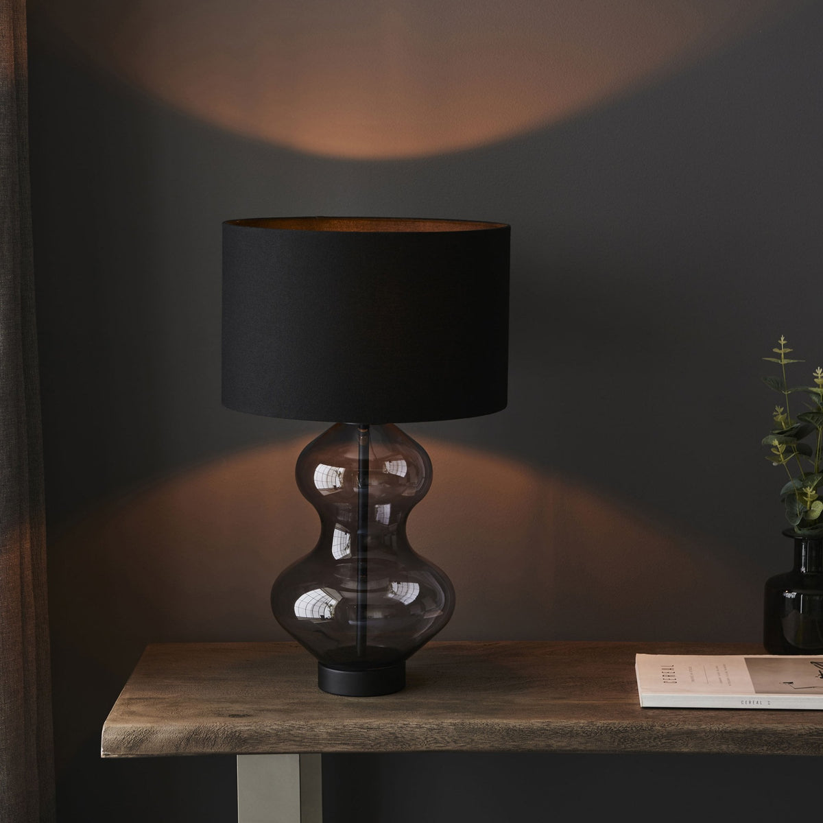 A modern table lamp with a dark gray hourglass-shaped glass base and a black cylindrical lampshade casting a warm glow.