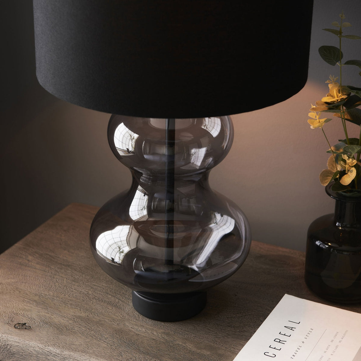 A close-up of a modern table lamp with a dark gray hourglass-shaped glass base and a black cylindrical lampshade.