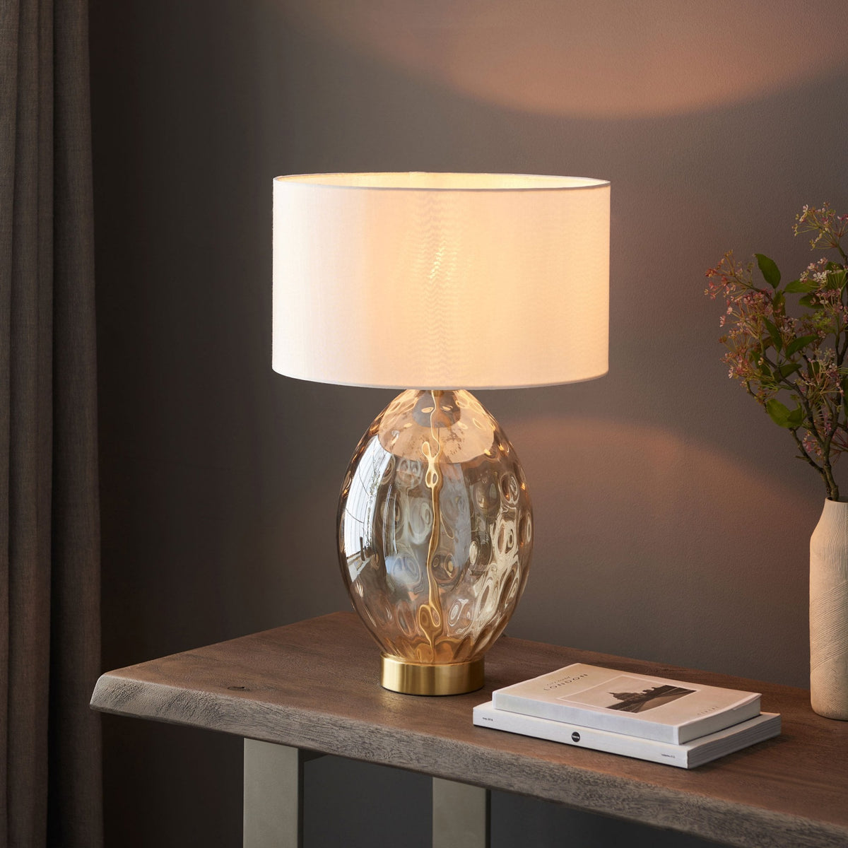 The Dimpled Glass Table Lamp next to a window, on top of a wooden table.