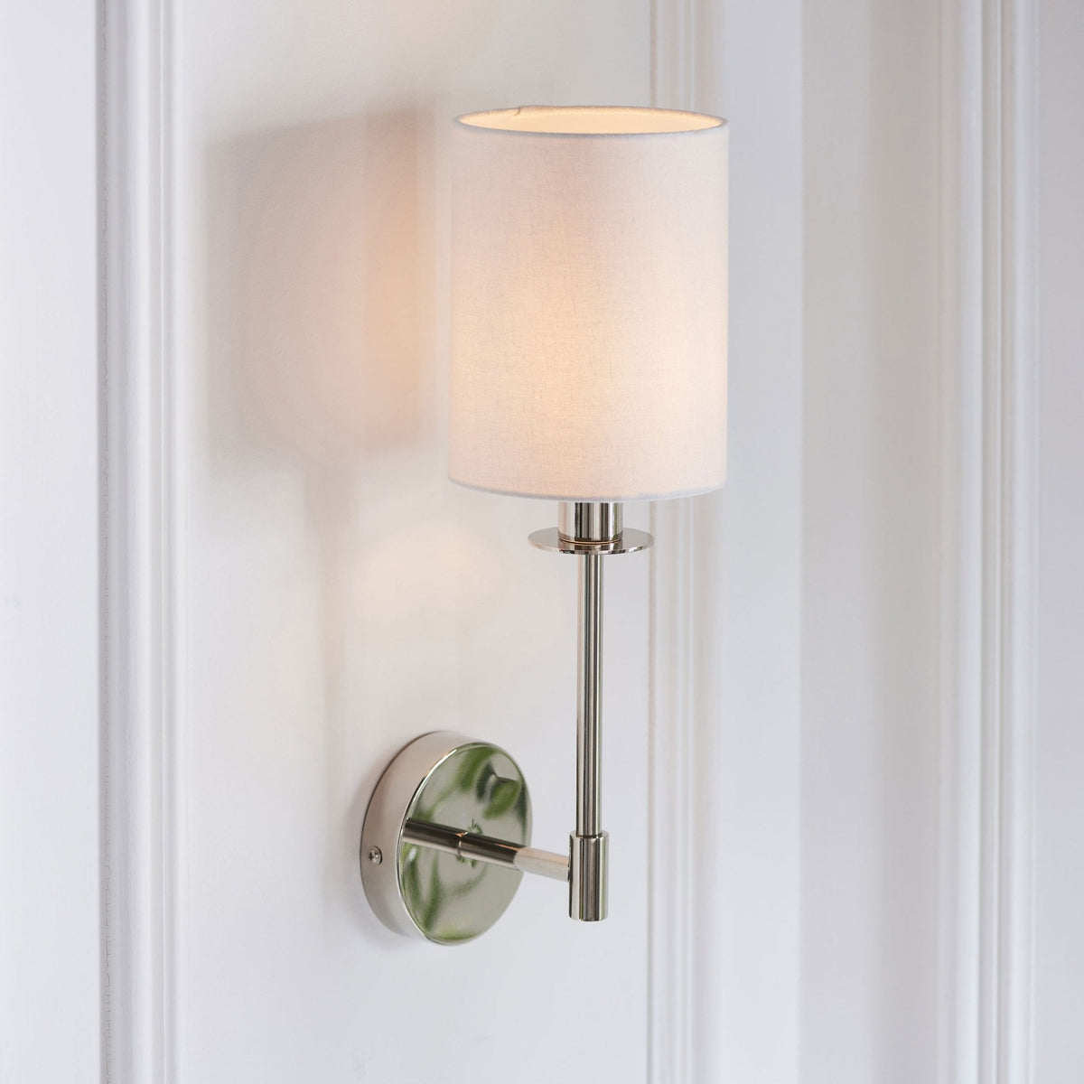 Polished nickel wall sconce with a white fabric drum shade, illuminating a white paneled wall.