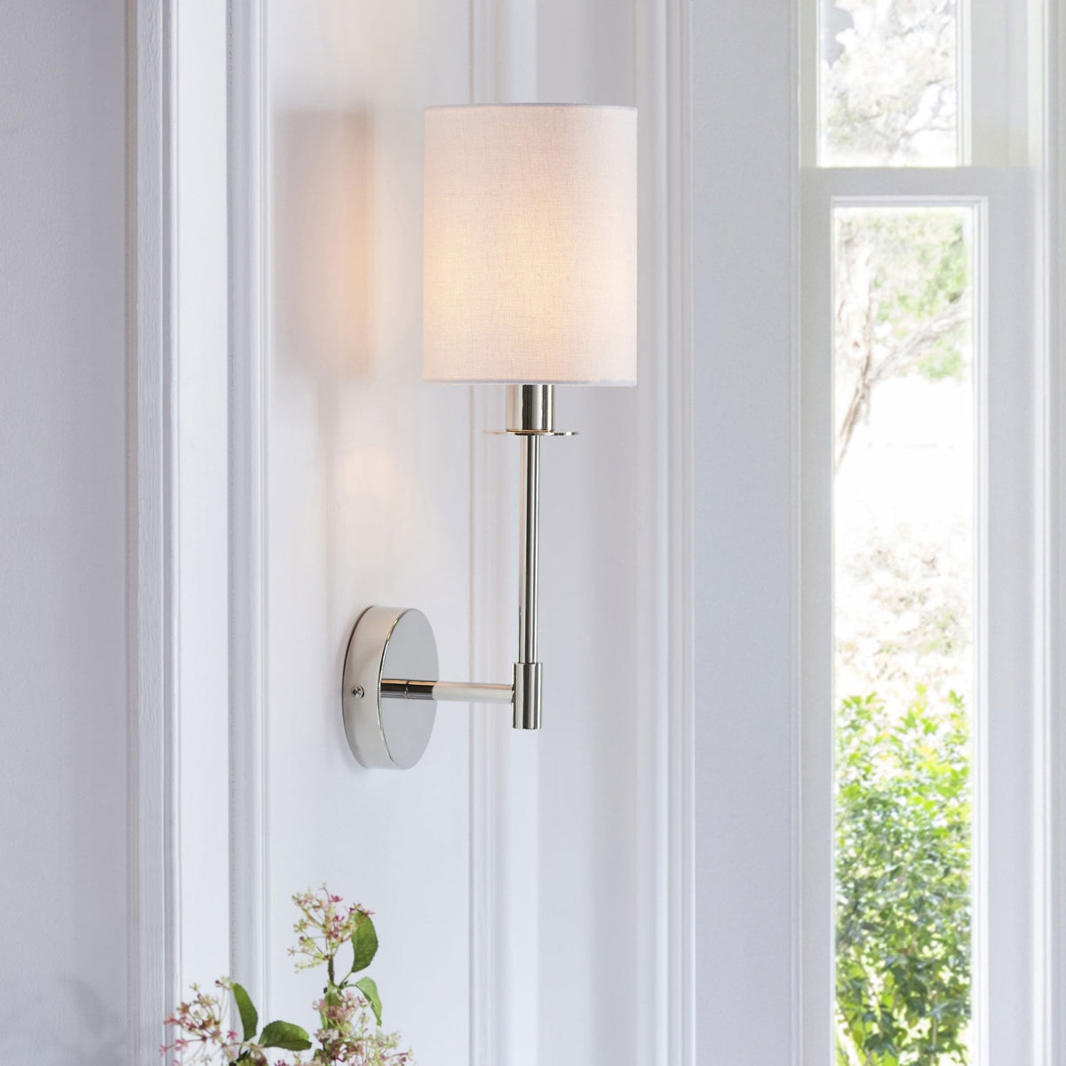 A polished nickel wall sconce with a white fabric drum shade, mounted on a white wall next to a window.