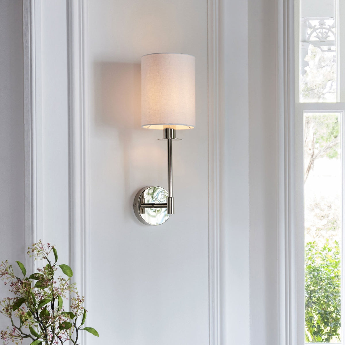 A polished nickel wall sconce with a white fabric drum shade, installed on a white wall by a window.