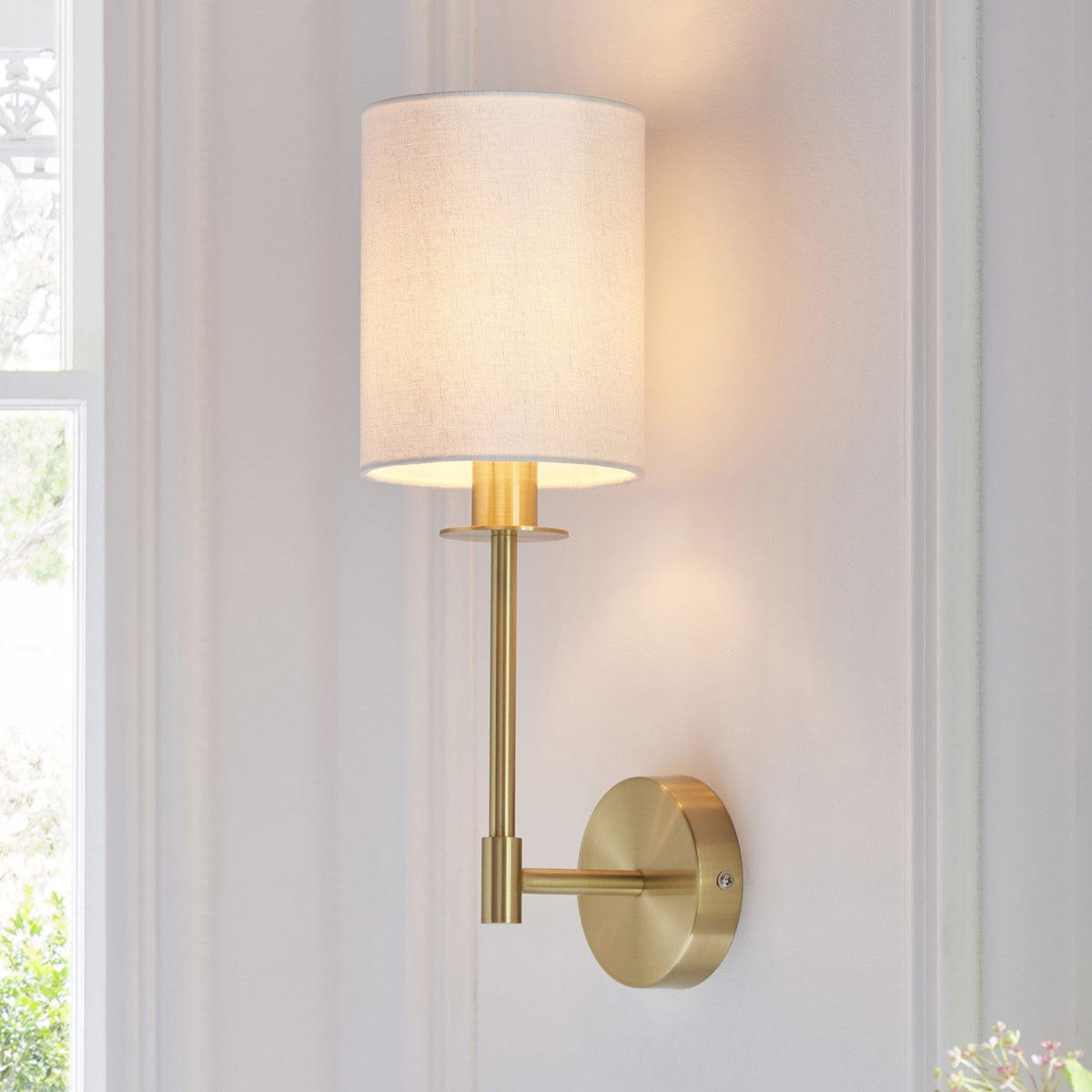 A modern brass wall sconce with a white fabric shade illuminates a light-colored wall next to a window.