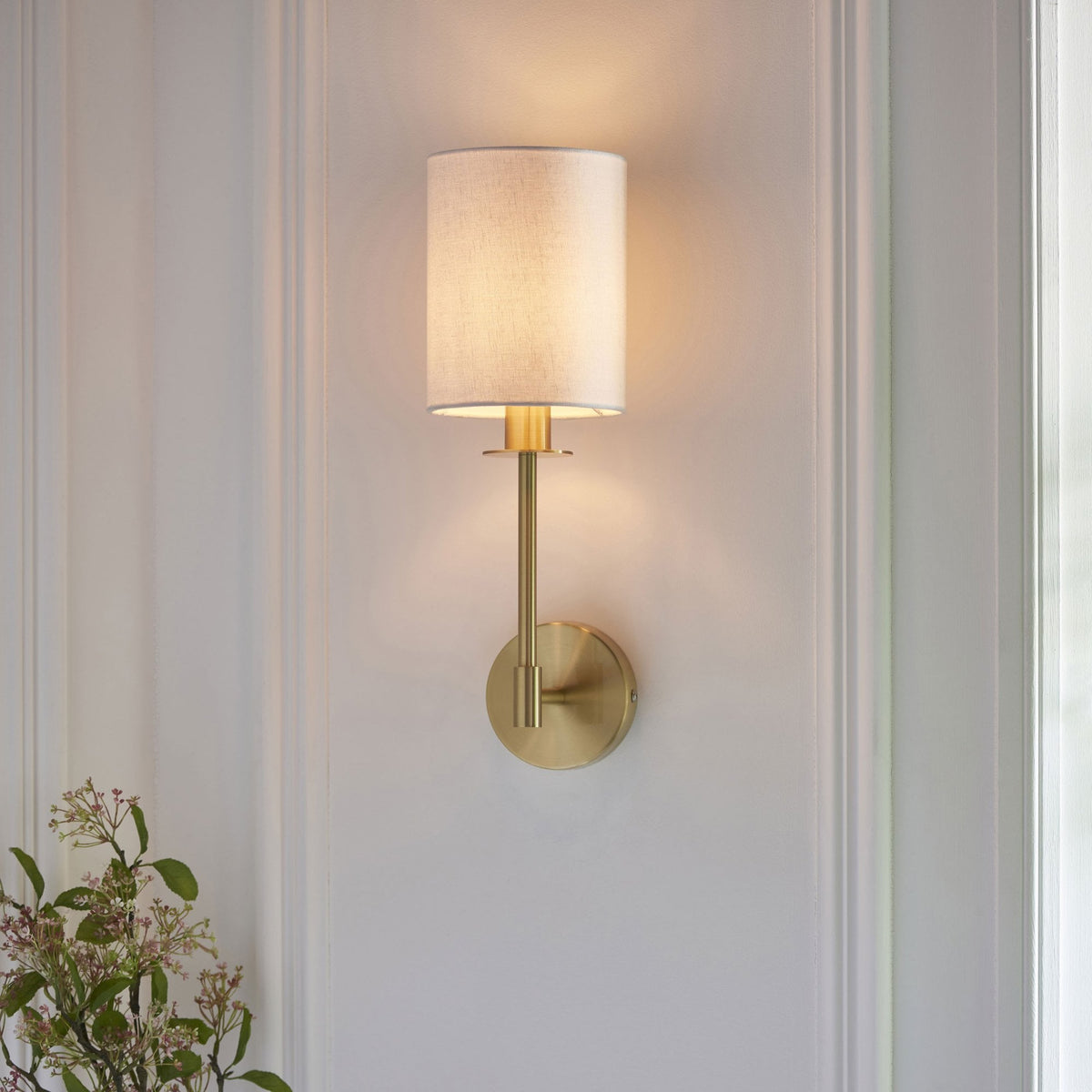 A chic brass wall light with a white fabric shade casts a warm glow on a paneled white wall above some flowers.