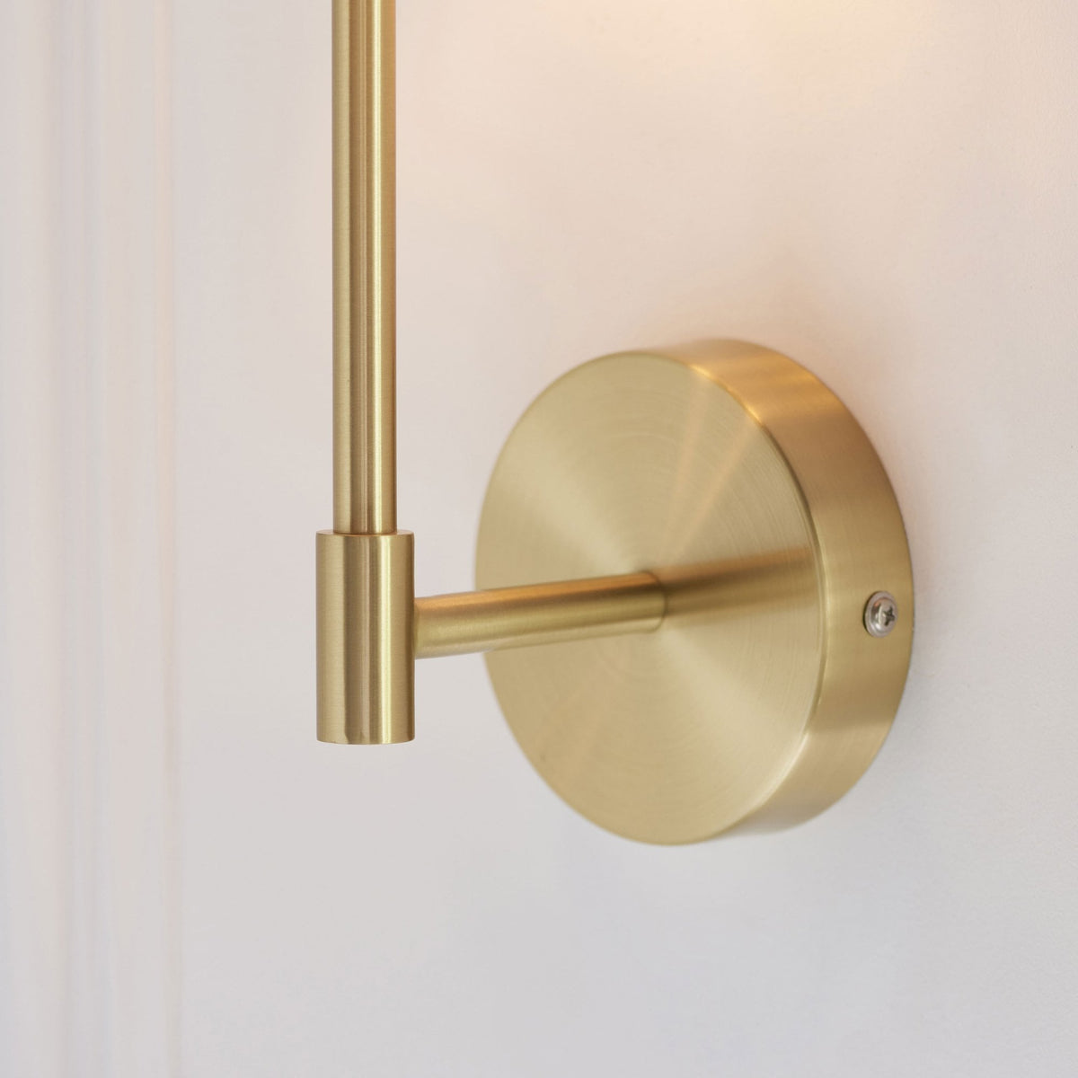 A close-up of the brushed brass base and arm of a wall-mounted light fixture against a white wall.