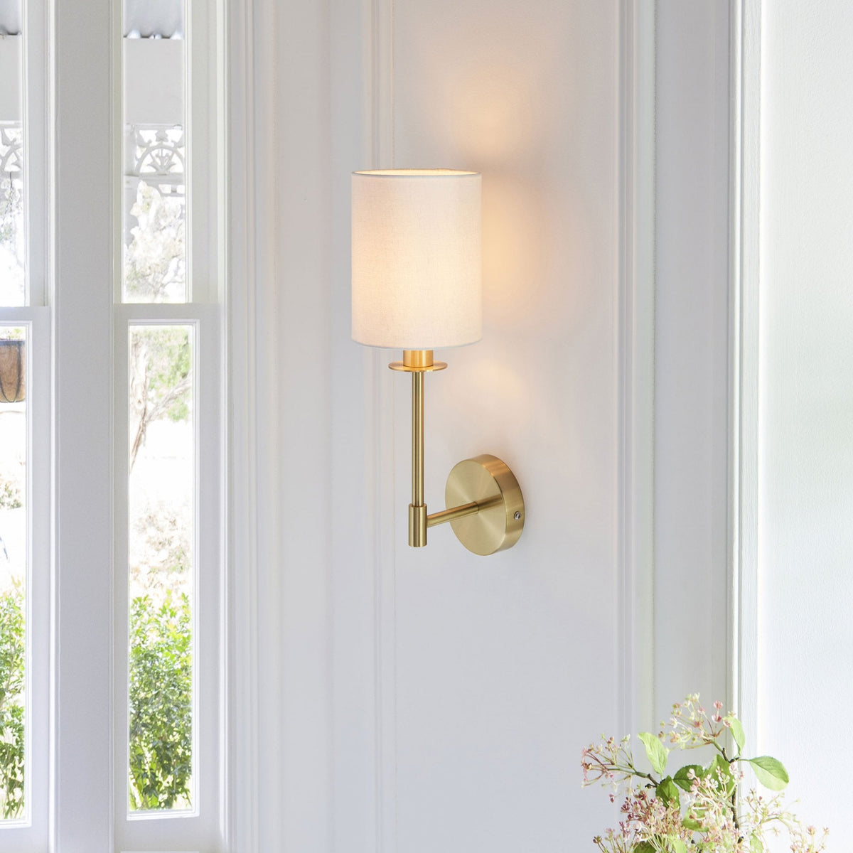 A sleek brass wall sconce with a white fabric shade lights up a wall next to a large window.