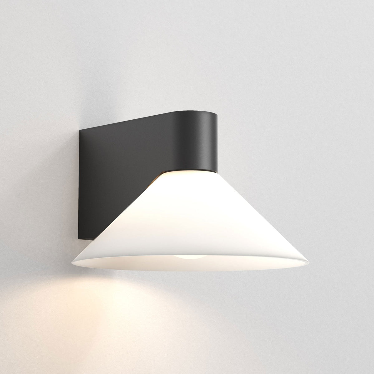 Black wall sconce with a white conical shade, casting a warm glow on a white wall