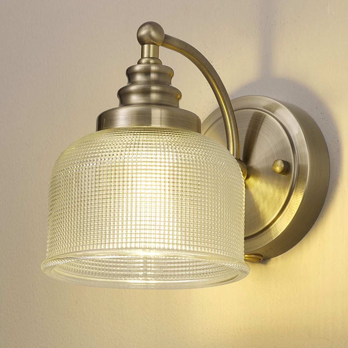 A brass wall light with a textured glass shade illuminates a warm glow against a grey wall.
