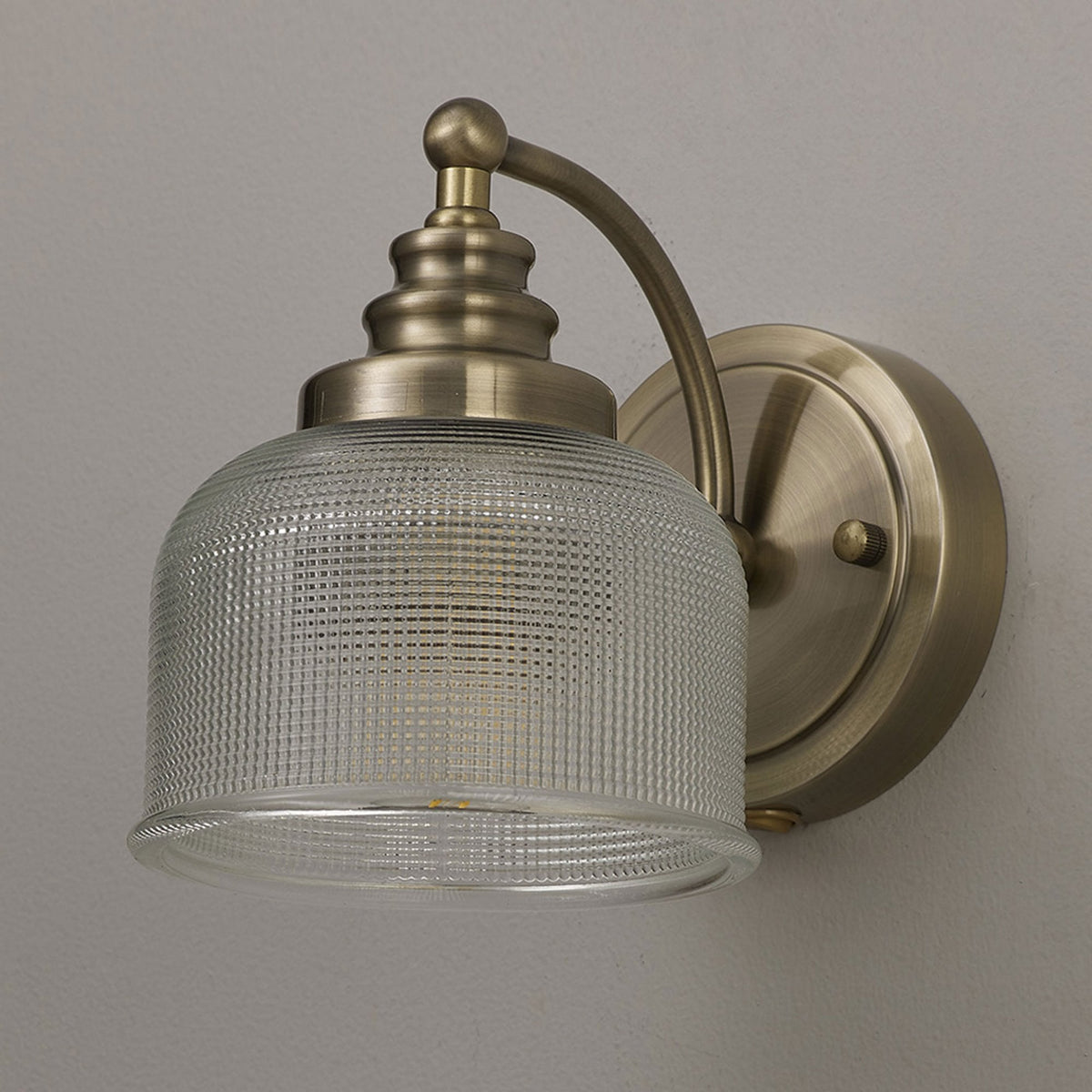 A brass wall light with a textured glass shade, unlit, mounted on a light grey wall.