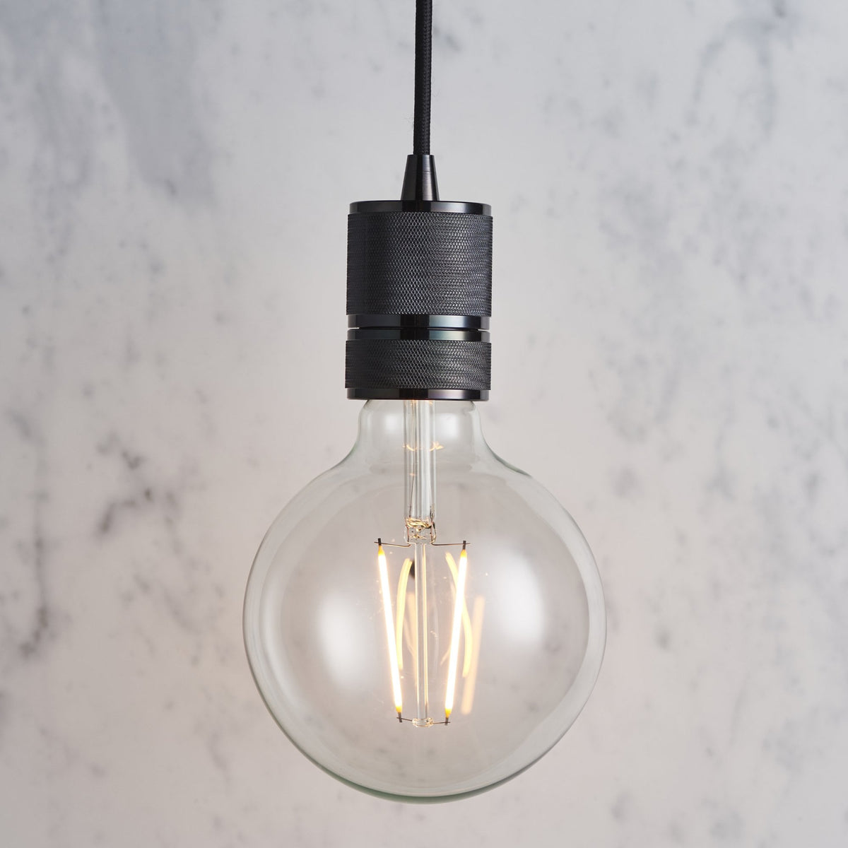 Close-up of a round clear bulb with visible filaments and a dark grey fixture against a marble wall.
