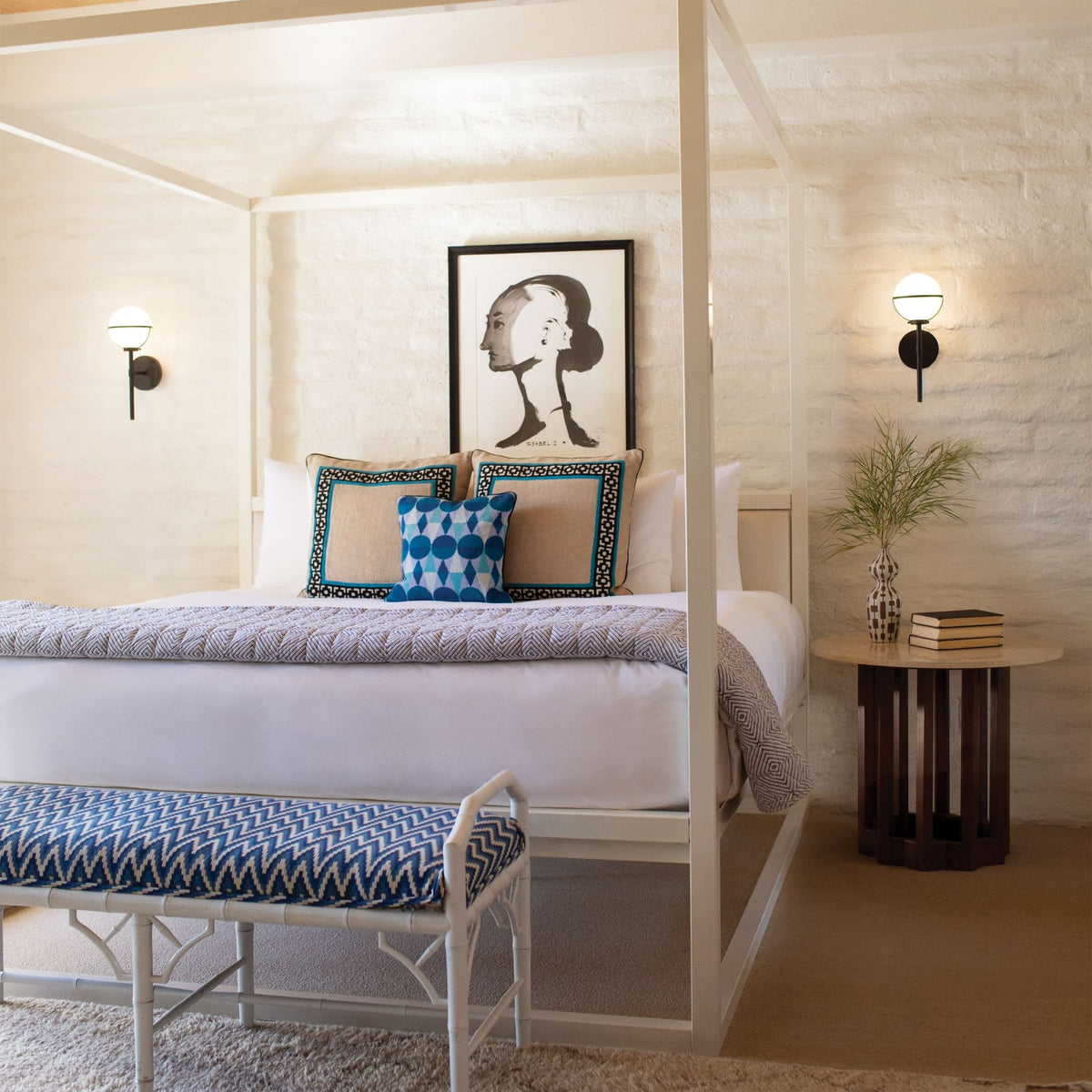 A bedroom with a white four-poster bed, blue and white patterned bench, and a framed abstract art piece