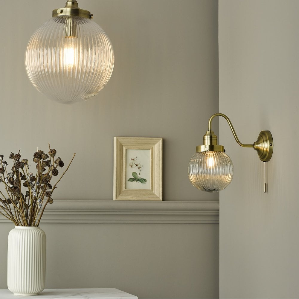 A brass pendant light and a brass wall light with ribbed glass shades illuminate a room with a framed botanical print.