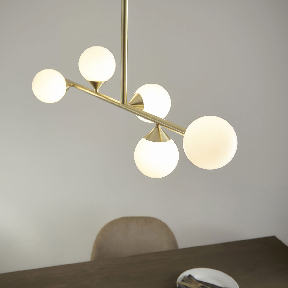 A close-up of a modern brass chandelier featuring five luminous globe lights over a dining table.