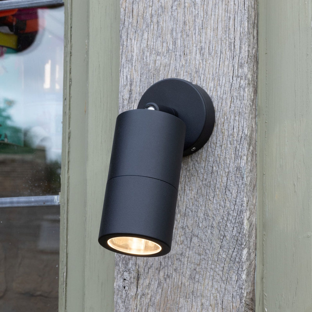 Black cylindrical outdoor light with a warm glow, mounted on a weathered wooden post next to a window.